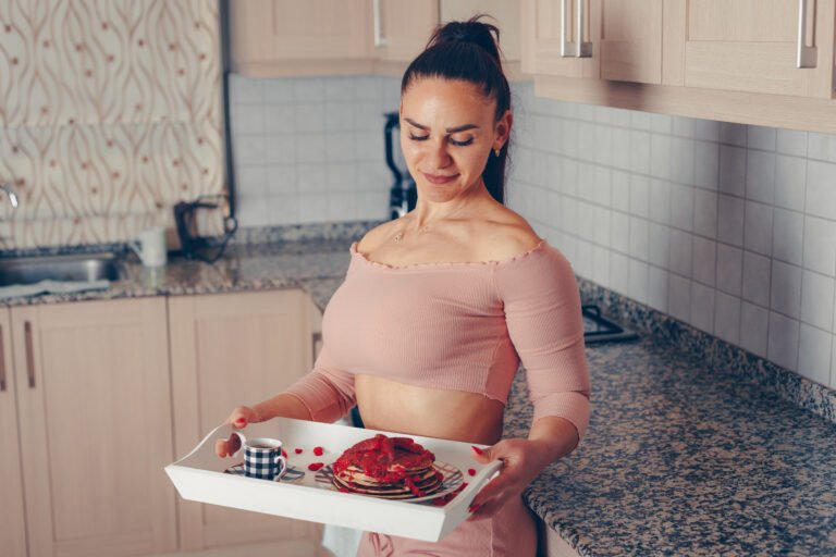 Attractive lady standing and holding meals in kitchen in salmon pink crop top and pants